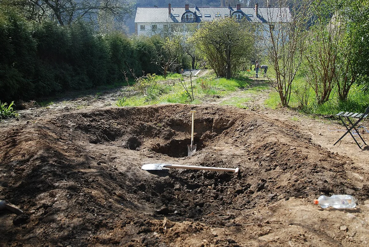 Een pas uitgegraven vijverkuil in donkere grond met een schep in de aarde en de uitgegraven grond op een hoop ernaast.