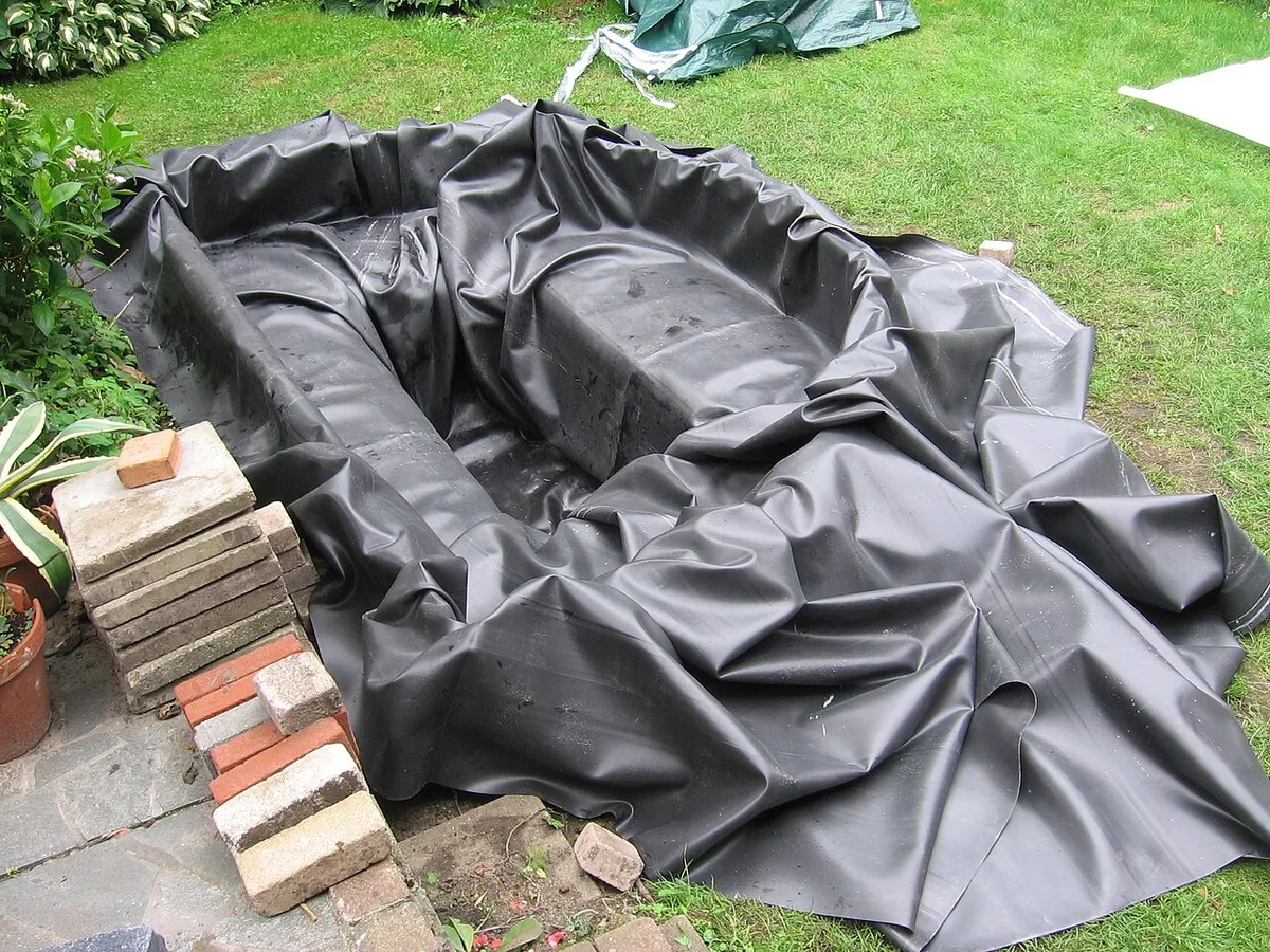 Zwarte vijverfolie ligt los uitgevouwen in een uitgegraven vijverkuil in een tuin, met een stapel stenen ernaast.