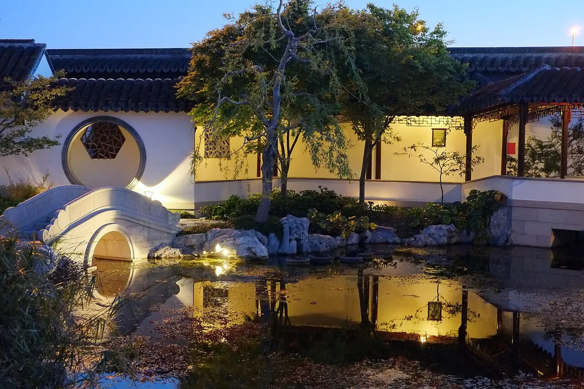 Een tuinvijver in de avond met warm verlichte beplanting en een boogbrug die in het stille water weerspiegelt.